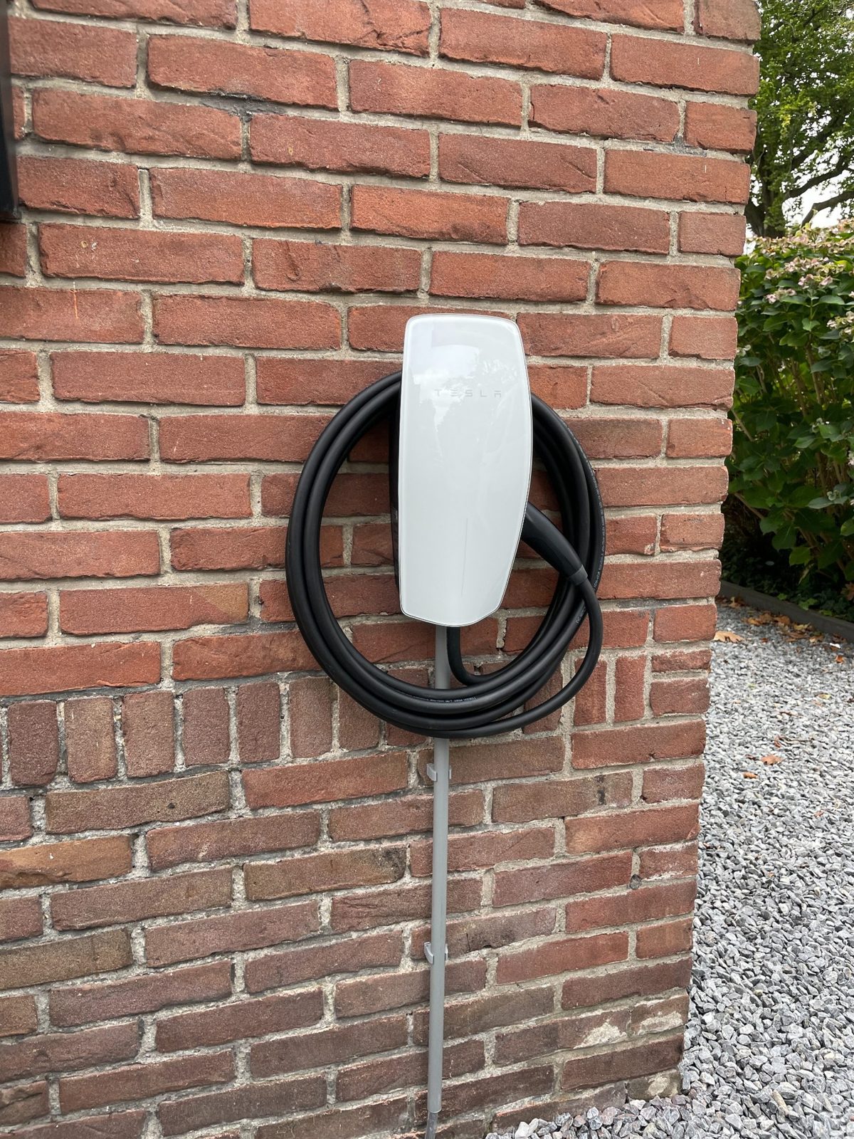 Tesla laadpaal met zwarte kabel bevestigd aan bakstenen muur naast grindpad