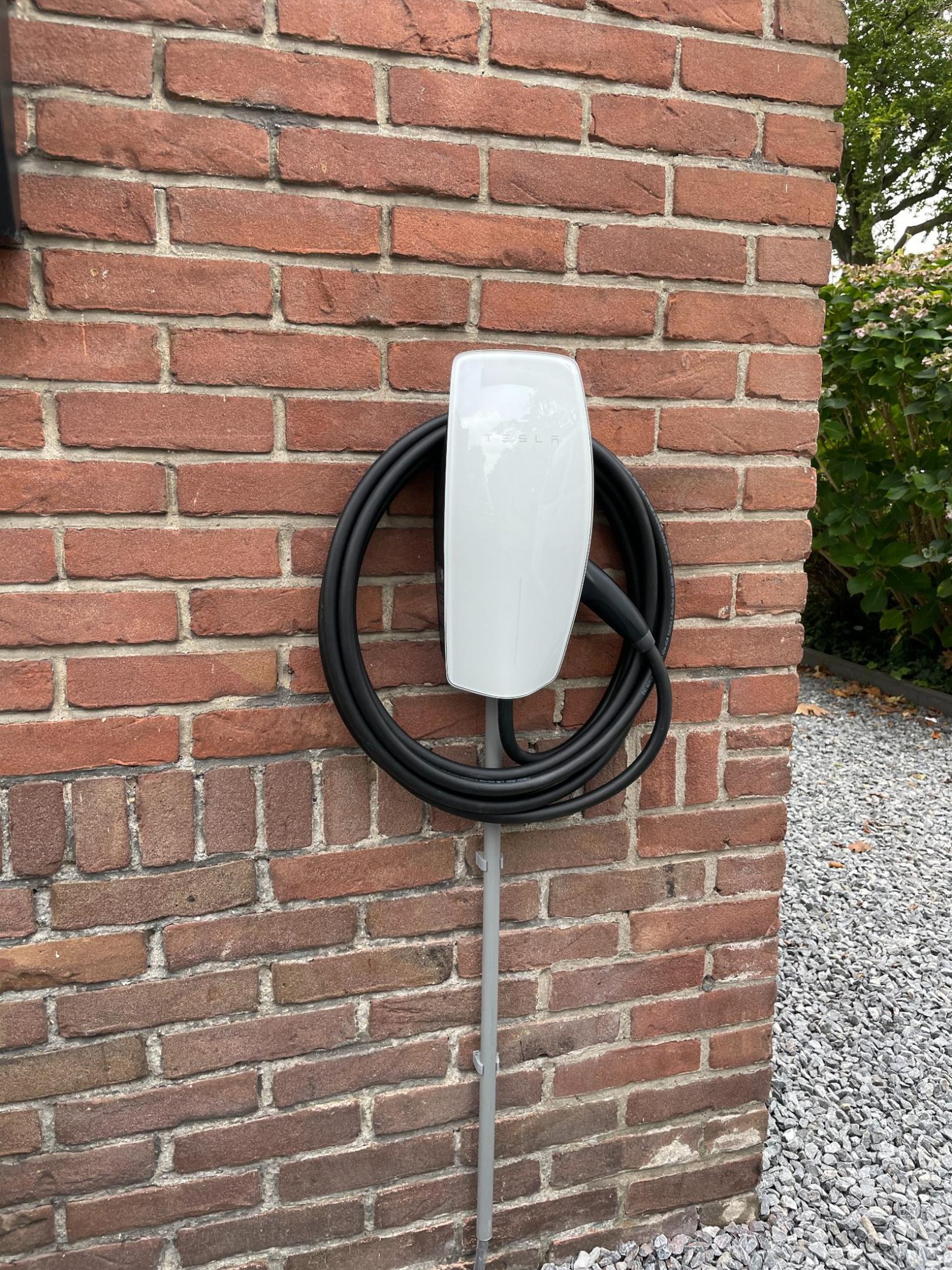 Tesla laadpaal met zwarte kabel bevestigd aan bakstenen muur naast grindpad